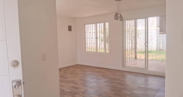 Living comedor visto desde la entrada. Piso vinílico, muros blancos, lámpara central y estufa empotrada a gas.